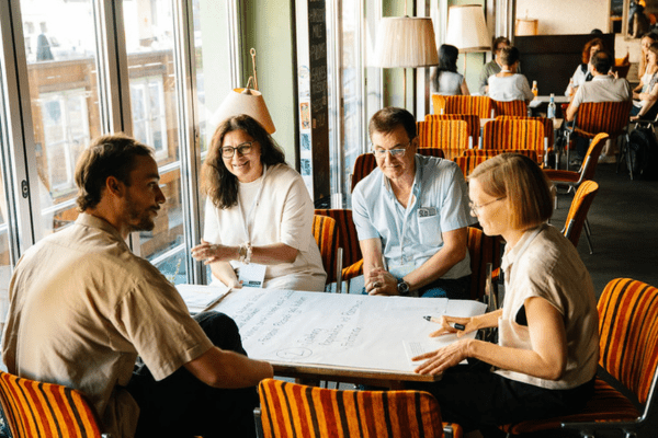 An einem Tisch sitzen in Café-Atmosphäre zwei Frauen und zwei Männer. Auf dem Tisch liegt ein Flipcahrt-Plakat. Die Gruppe diskutiert angeregt. Im Hintergrund arbeitet eine weitere Gruppe.