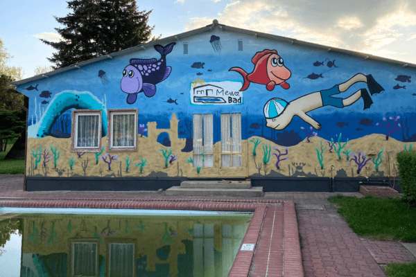 Das Foto zeigt das Becken und das Graffiti Fassade der Baracke. Das Graffiti zeigt eine Unterwasserwelt und einen Taucher, der zwischen Fischen und Korallen schwimmt.