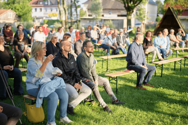 Das Foto zeigt Menschen, die auf Bierbänken sitzen und in eine Richtung schauen. Weciehs Abendlich fällt auf die Gruppe.