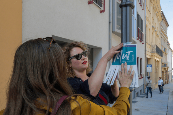 Das Foto zeigt zwei Frauen, die ein Plakat für Mut zum Mitnehmen an einen Laternenmast kleben.