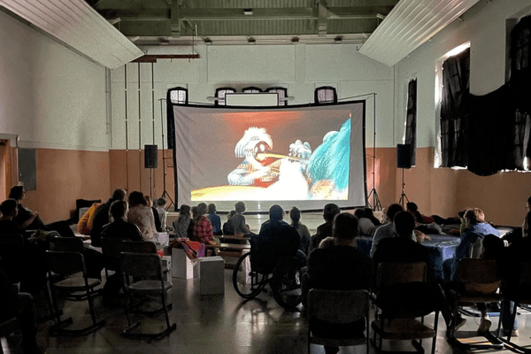 Das Foto zeigt eine Kinoveranstaltung in der alten Turnhalle: Menschen sitzen im Dunklen vor einer großen Leinwand.