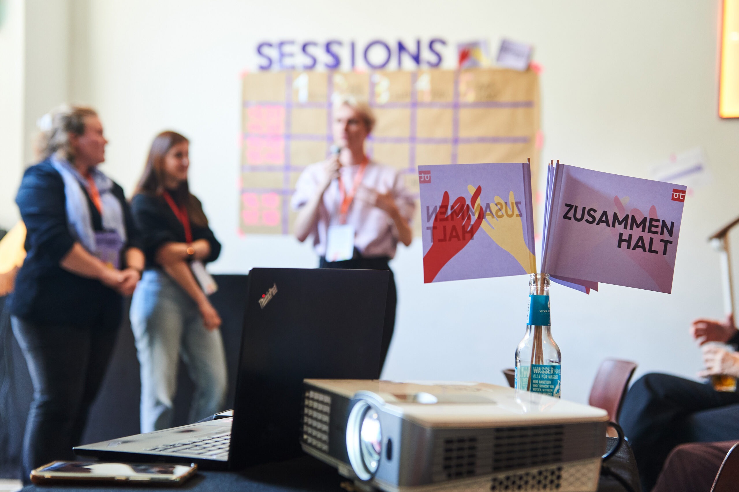 Auf zwei Fähnchen ist das Wort "Zusammenhalt" zu sehe. Im Hintergrund sind drei Personen, die vor einem Sessionplan stehen.