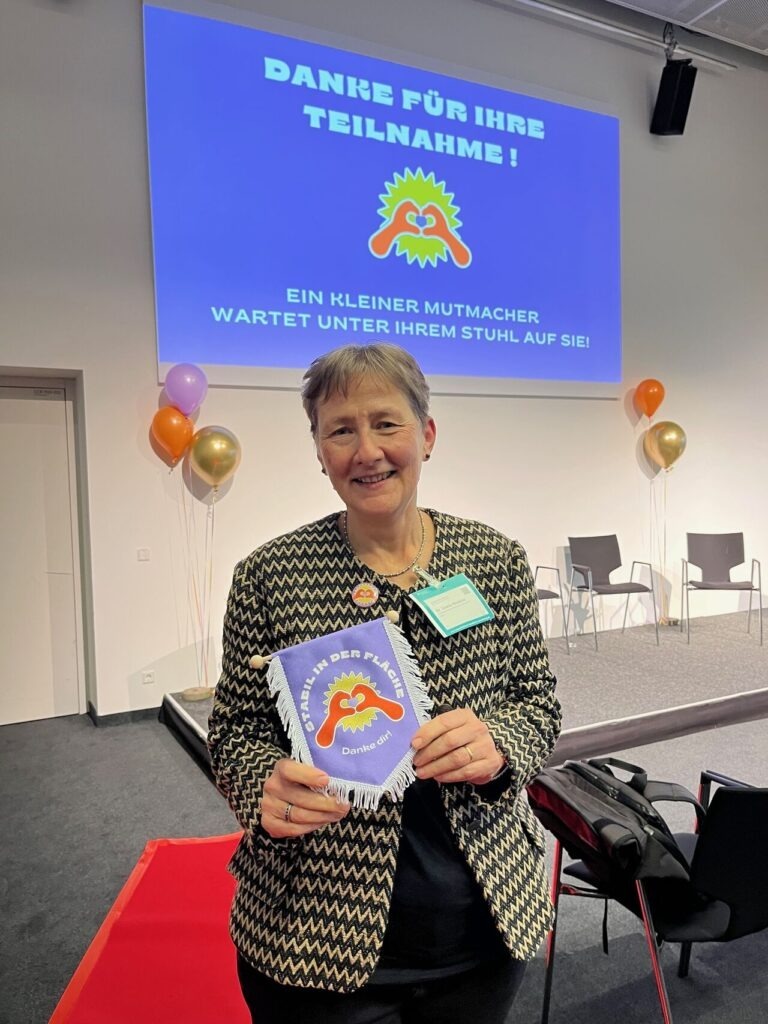 Eine Frau mit kurzen grauen Haaren blickt in die Kamera. Sie hält einen lila Wimpel in der Hand, auf dem zu lesen ist: Stabil in der Fläche. Danke dir!