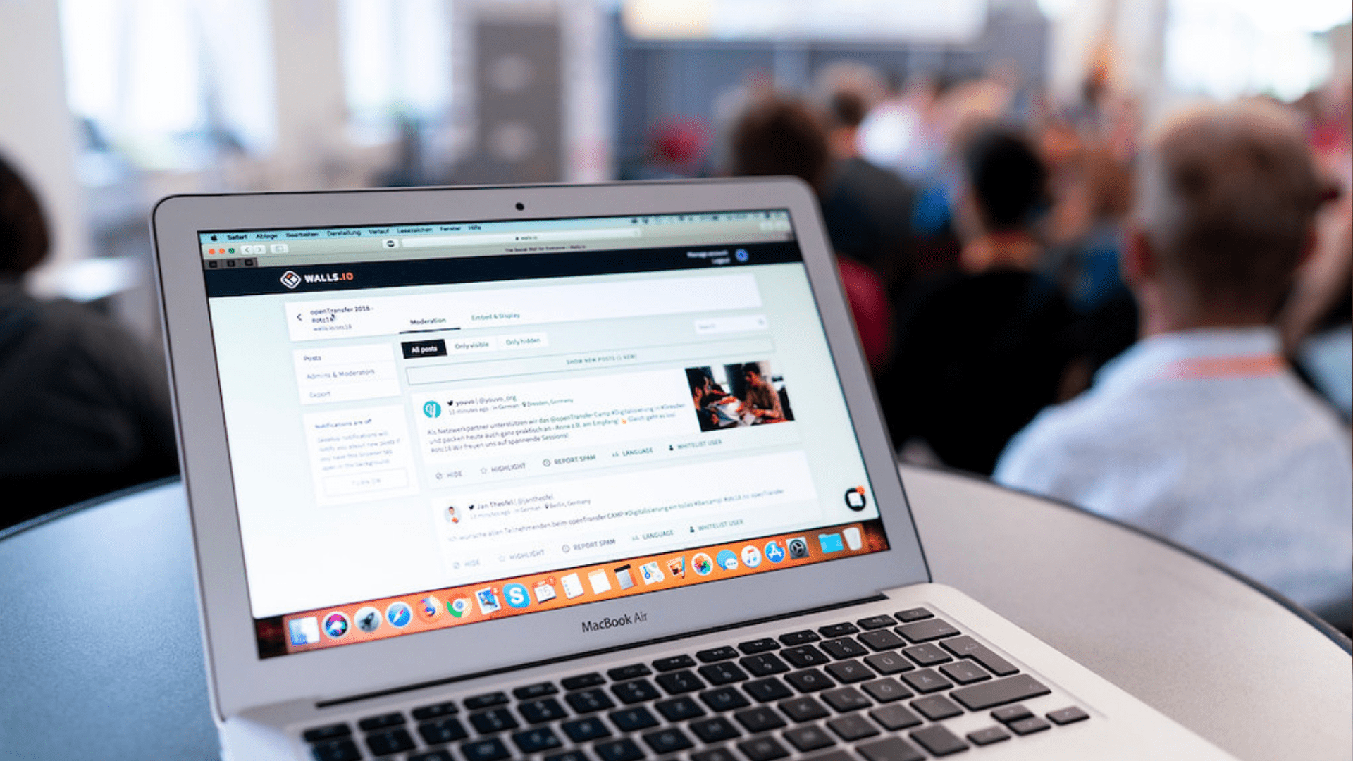 Ein aufgeklappter Laptop. Im Hintergrund ist unscharf ein Publikum zu erkennen.