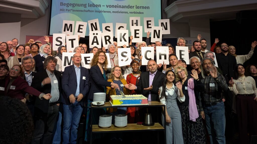 Ein Gruppenbild mit vielen Menschen. Sie halten Zettel mit Buchstaben in die Höhe, die zusammen ergeben: Menschen stärken Menschen
