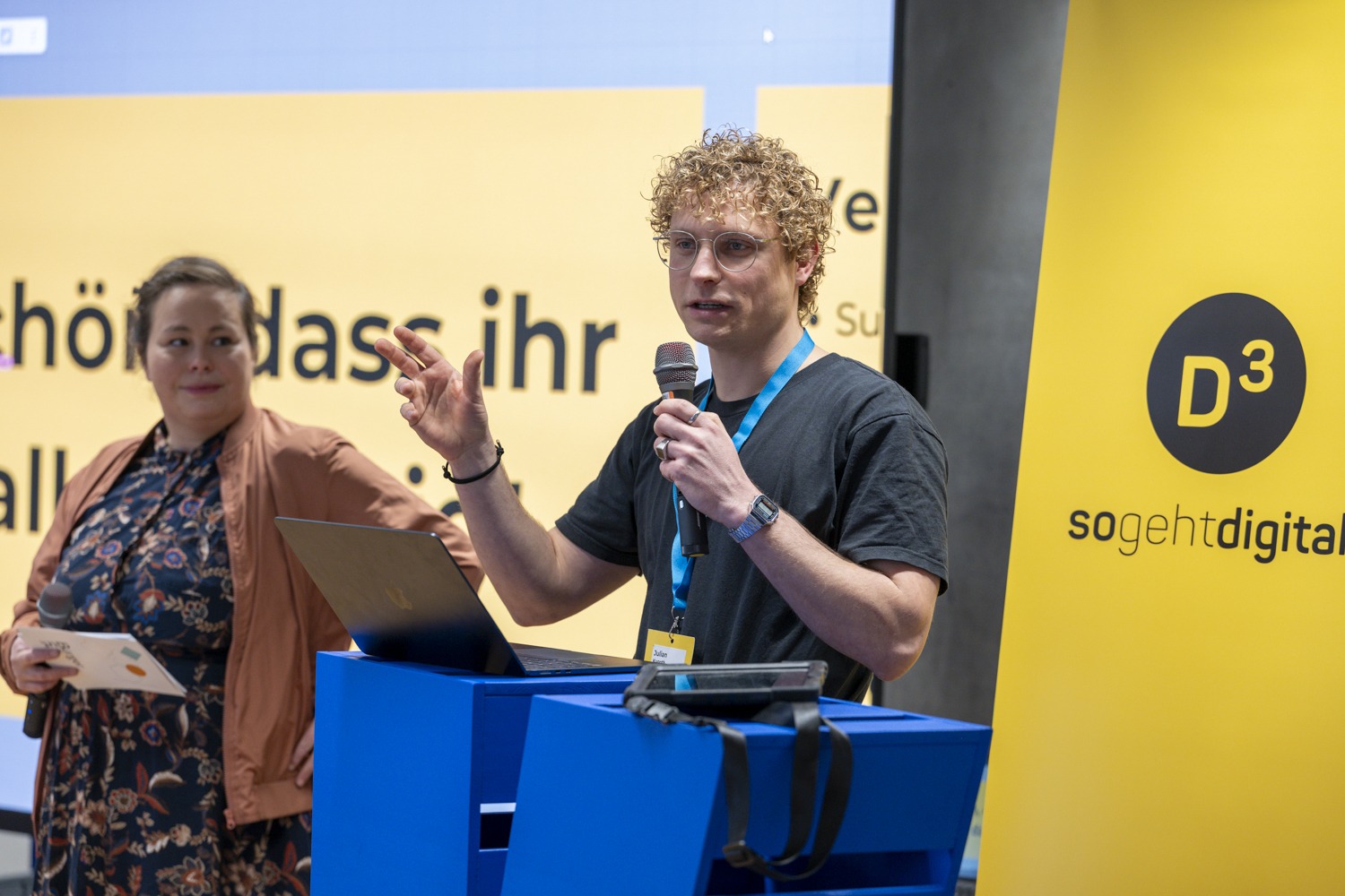 Ein blonder junger Mann spricht gestikulierend in ein Metro. Neben ihm steht eine Frau. Im Hintergrund ist ein Banner mit dem Logo des Programms D3 - so geht digital.